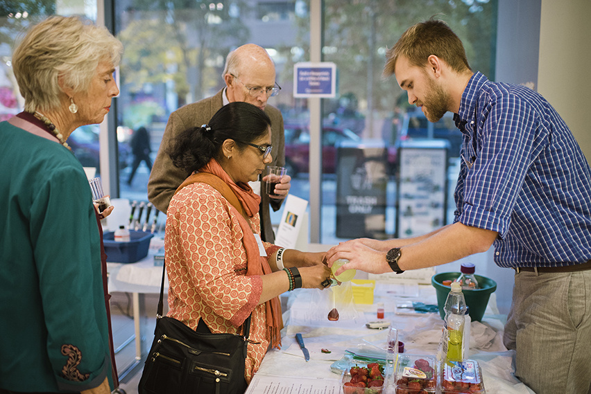 three people interact with a man giving a science demonstration involving strawberries