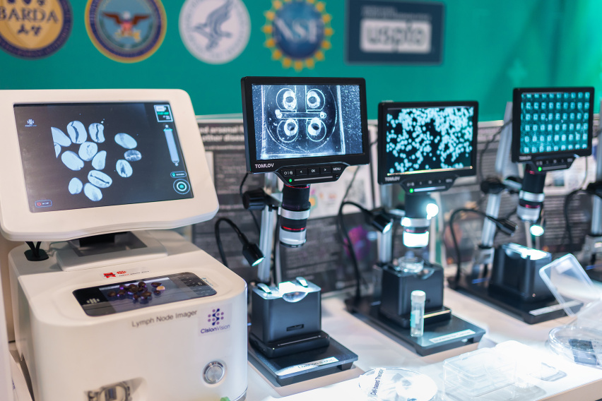 Several devices with view screens on a table, with the seals of various governmental organizations on a wall behind