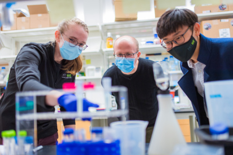 Michael Birnbaum and two students looking at an experiment on a lab bench covered in flasks and tubes