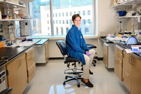 Julian Zulueta in a blue lab coat, seated on a rolling chair between two lab benches.